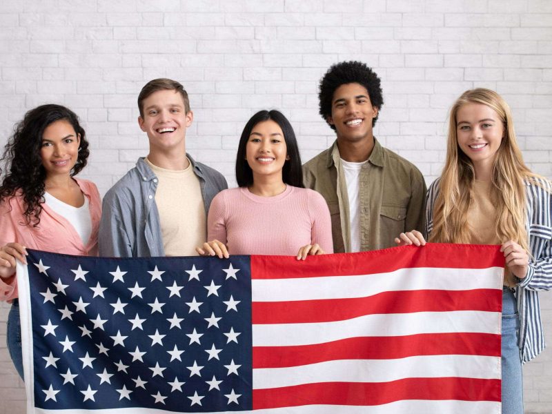 Immigration, relocation and study abroad. Cheerful young international students hold USA flag, looking at camera and ready to learn, on white brick wall background, copy space, studio shot
