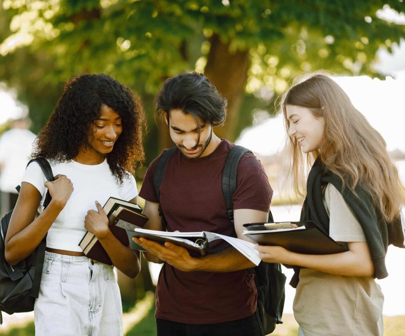 Group of international students standing together in park at university. African and caucasian girls and indian boy talking outdoors. Three friends looking in a book.